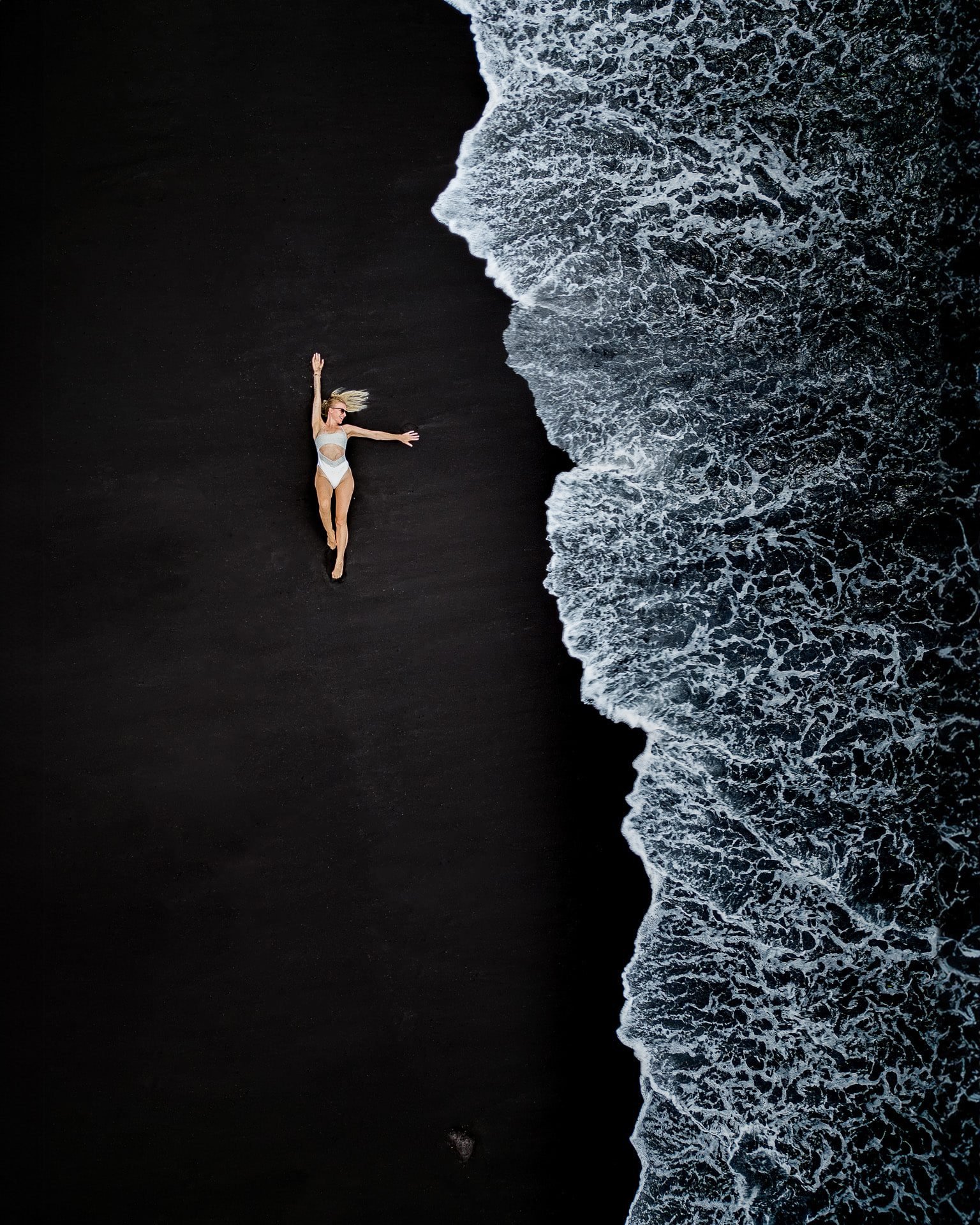 Person am schwarzen Strand, die Brandung von oben