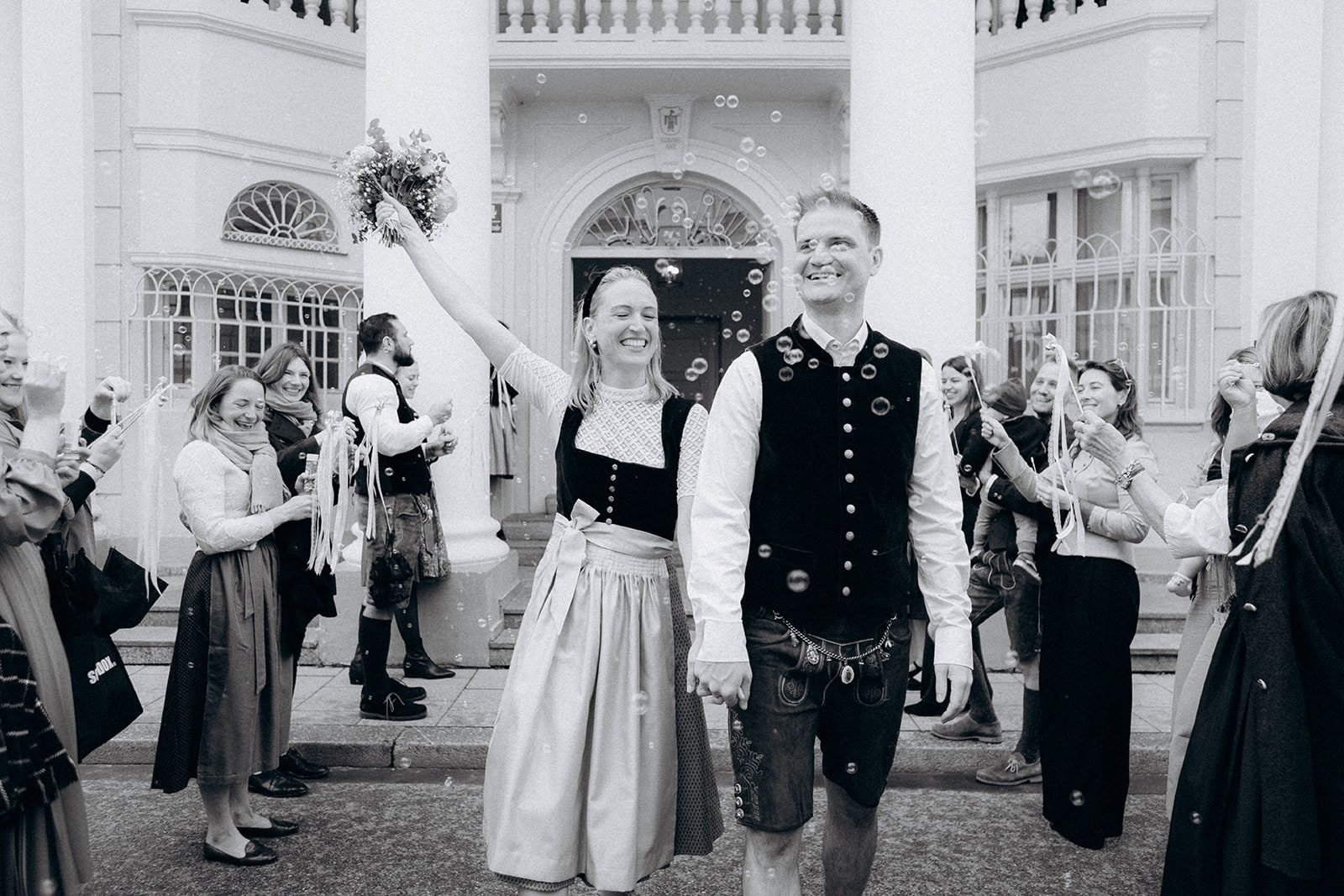 Brautpaar in Tracht läuft lachend durch ein Seifenblasen-Spalier der Gäste, Schwarzweiß